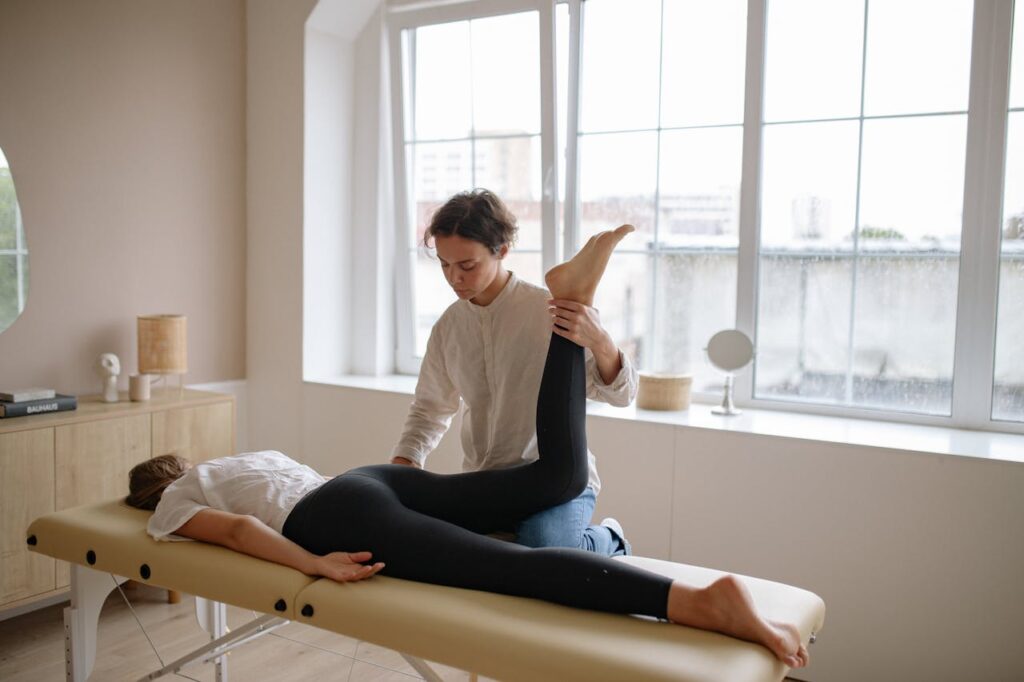 Massage therapist provides leg therapy to client on a table in a sunlit room.